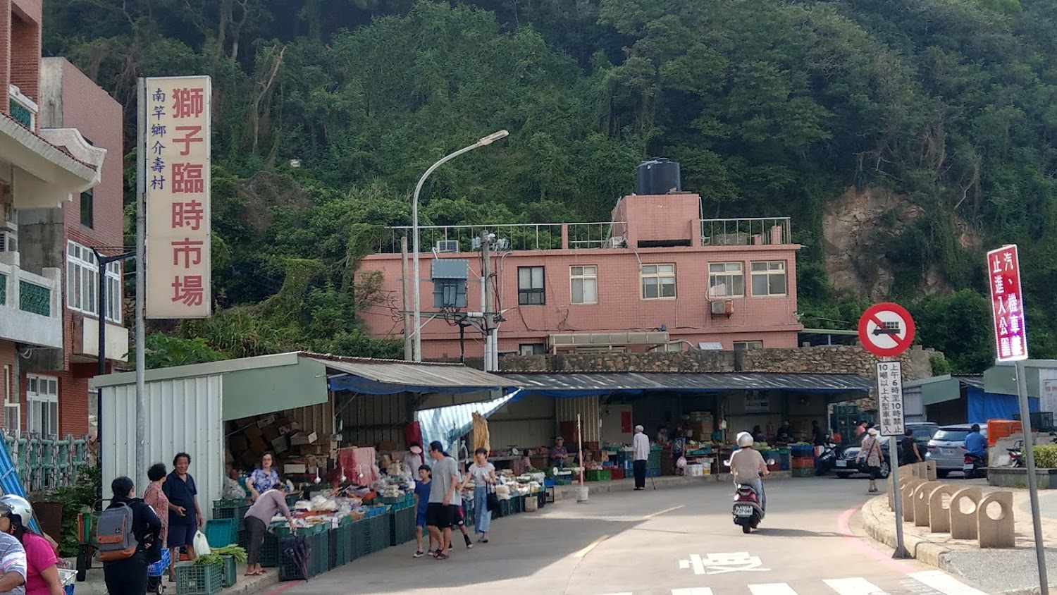 這張圖片主要是獅子臨時市場的場景，有攤販販賣各式蔬果與日用品。市場前來往的行人及機車顯示出當地的生活日常，背景有住宅和綠樹山坡。