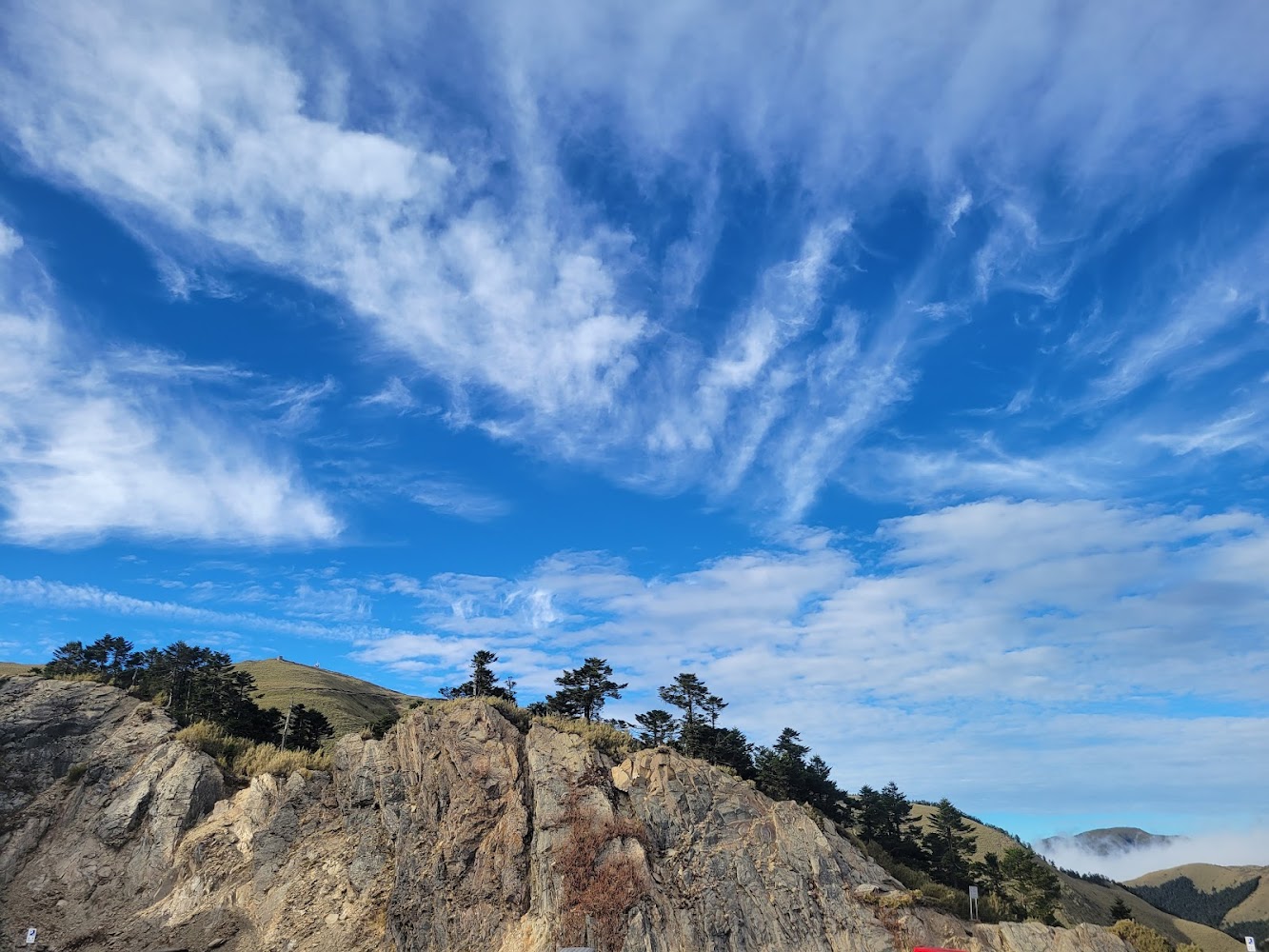 這張圖片主要呈現藍天、大片白色雲朵與壯麗的岩石山丘,山丘上有零星叢林樹木。整個景緻典雅自然,無浮水印。