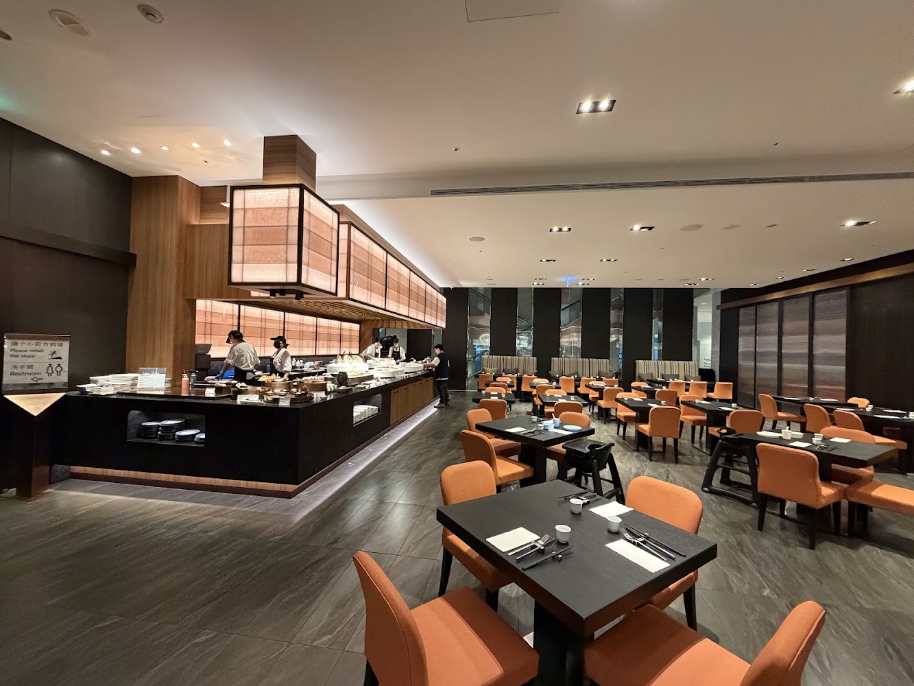 這張圖片展示了一家現代風格的自助餐廳,內有整齊排列的橙色座椅與深色餐桌,左側有開放式自助餐檯及工作人員,環境明亮整潔,圖片左方有洗手間指標看板,無明顯浮水印。