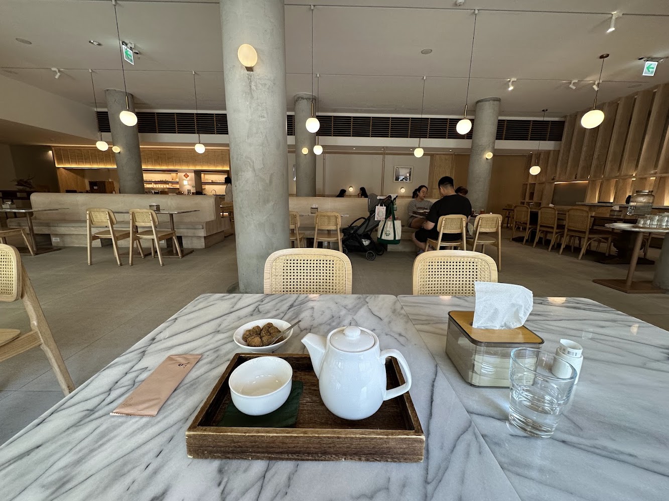 這張圖片顯示一家現代風格的咖啡館或餐廳，桌上有茶壺、杯子、點心、紙巾盒和水杯，環境明亮舒適，擺設簡約，有顧客在用餐。