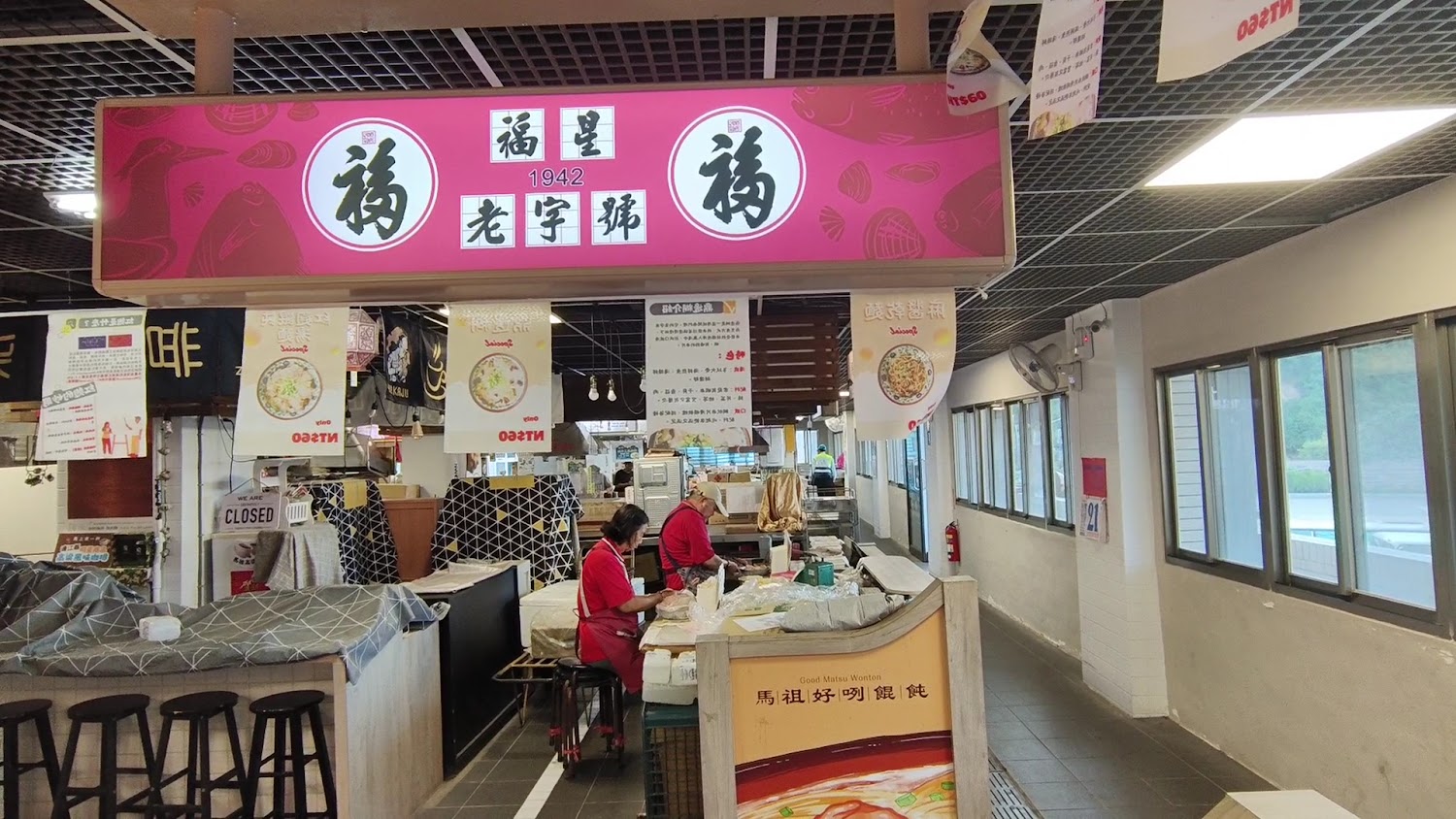 這張圖片展示一家名為福星老字號的餐飲攤位，兩位店員正在工作，現場主要賣餛飩麵食，攤位乾淨明亮，有菜單與美食宣傳布條裝飾。