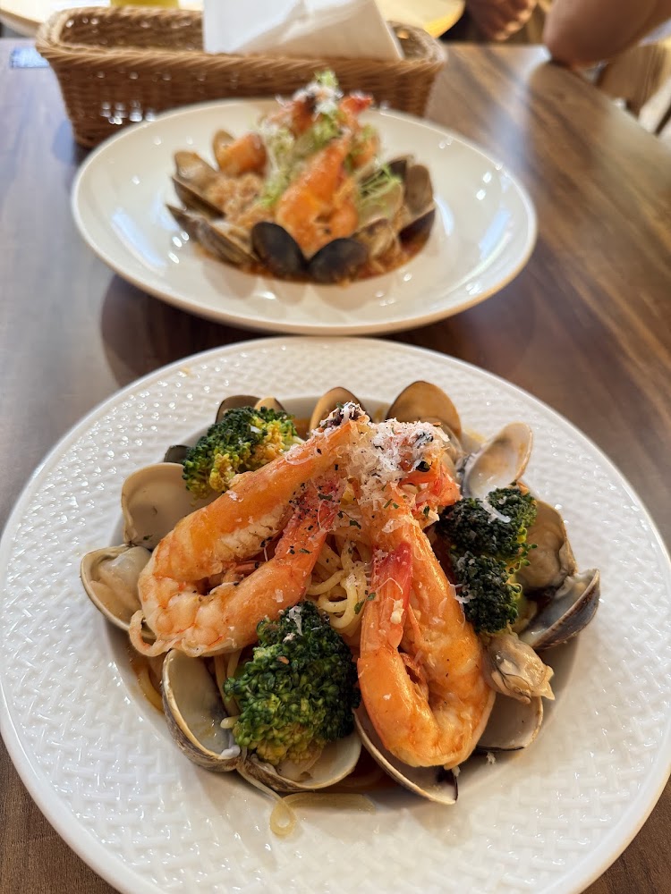圖片中有一道豐富的海鮮義大利麵，以蝦子、蛤蜊和花椰菜為主食材，擺盤精美，後方還有另一盤相似的海鮮料理。