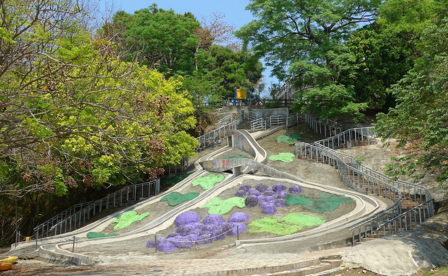 這張圖片主要是一座戶外滑梯遊樂區,滑梯蜿蜒於山坡上並有紫色與綠色的造景裝飾,兩側設有扶手步道供人上下,周圍被茂密樹木圍繞,無明顯浮水印。