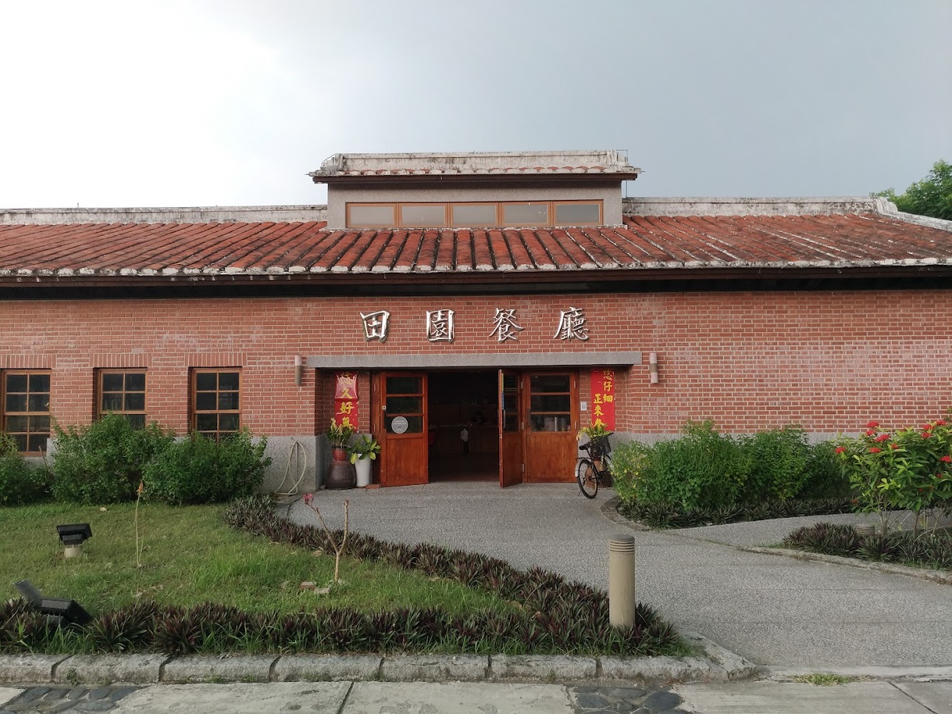 這張圖片主要是一棟紅磚瓦屋的建築，正面掛有『田園餐廳』的招牌，門口擺放著盆栽和腳踏車，周圍有綠色植物與花草，環境清幽。