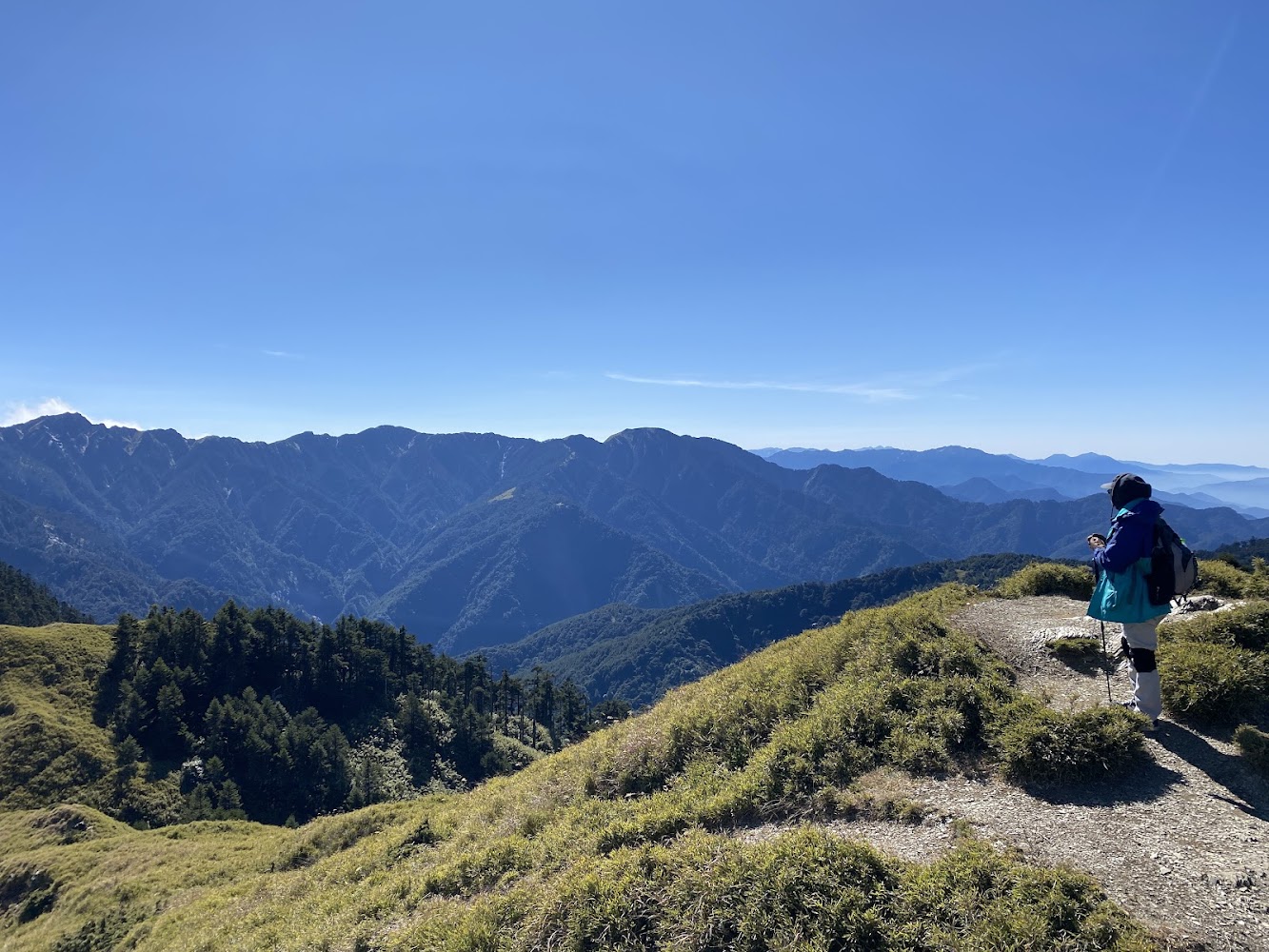 圖片中主要呈現一位登山者站在山徑上,遠眺壯麗的山脈景色,天空晴朗,藍天與層層山巒構成開闊的自然景觀,圖片無明顯浮水印。