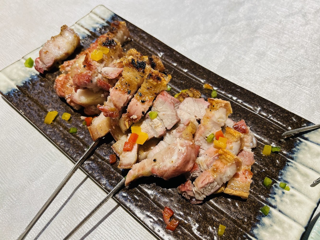 厚切烤豬肉串擺在長方形陶瓷盤中，上面點綴彩色蔬菜丁