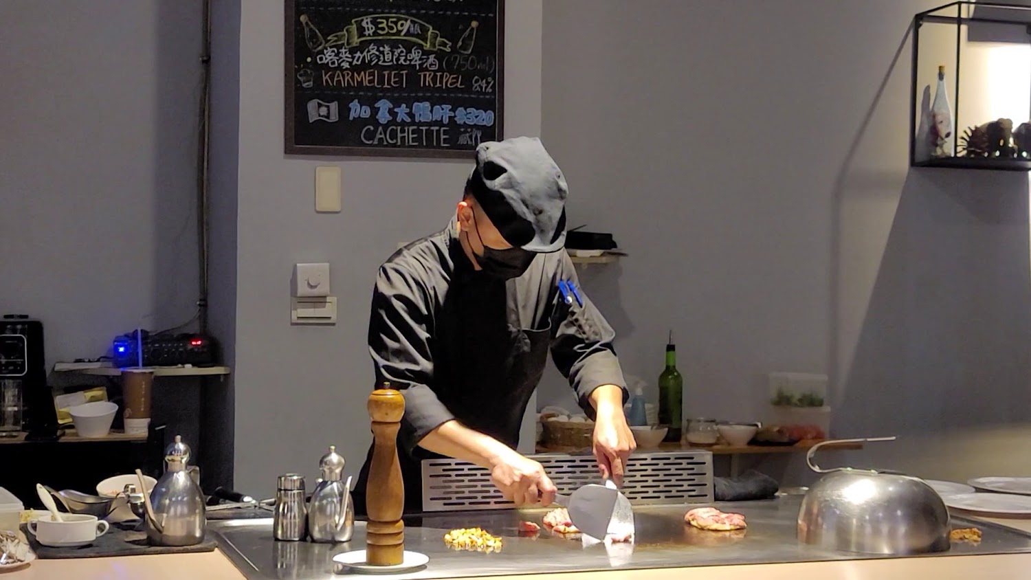 這張圖片主要是一位廚師在鐵板燒台前烹飪食材，周圍有各式調味品器皿，以及一些廚具和食材，背景有寫有菜單的黑板。