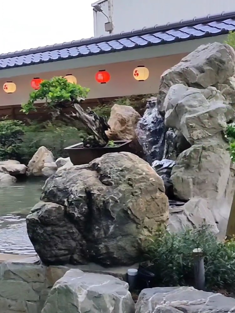 日式庭園，有大型石頭、流水、池塘與綠植，背景有紅色燈籠與傳統屋簷。