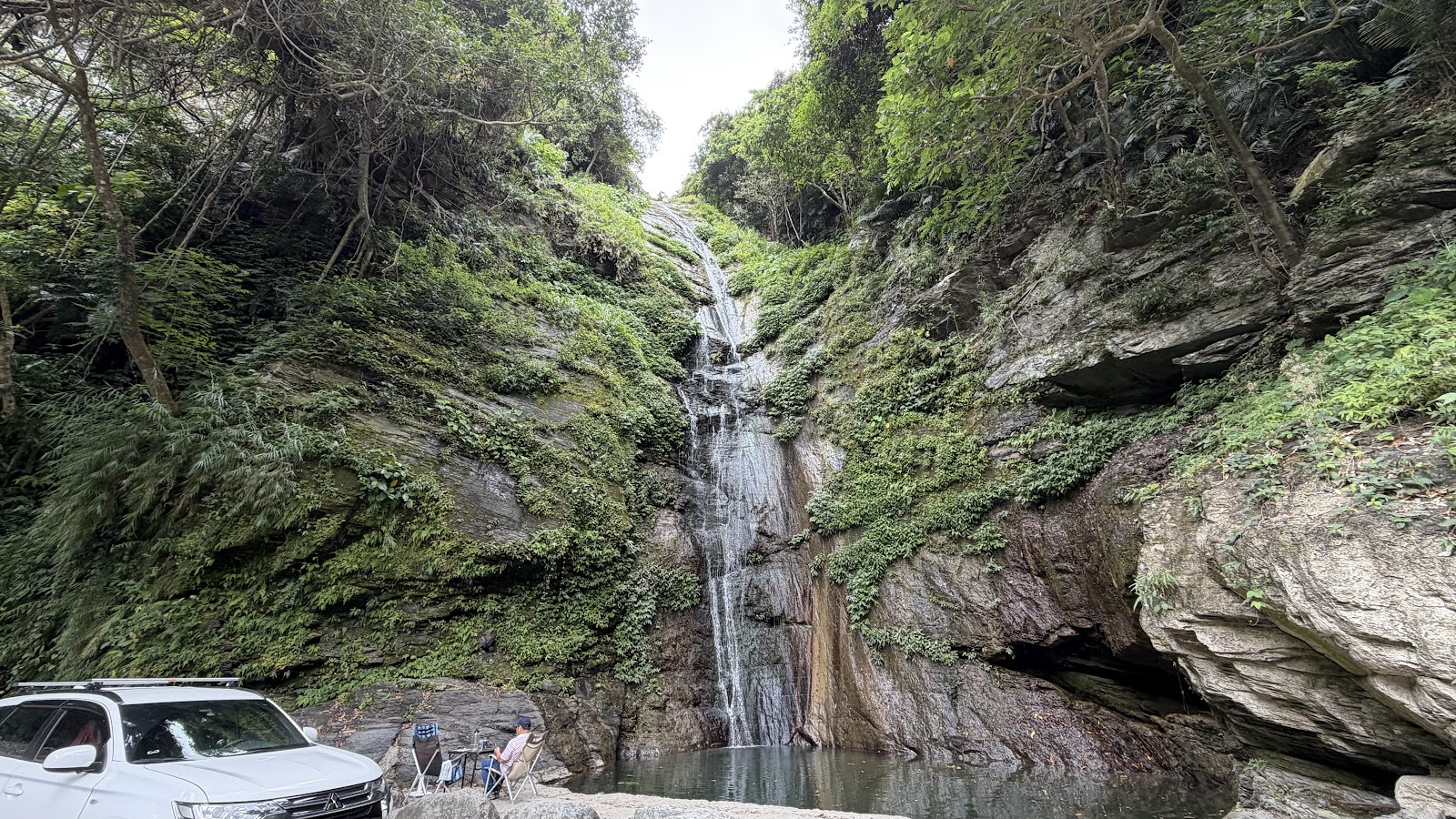 知本林道白龍瀑布全景，停有白色SUV車輛，兩位遊客席地休憩，岩壁兩側綠意盎然