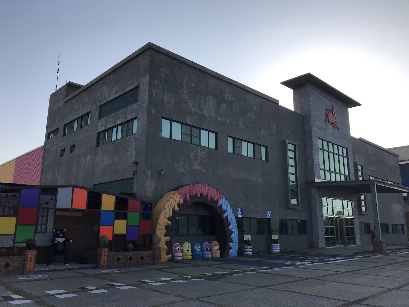 現代工業風格的建築物，外觀以灰色為主，前方有彩色裝飾和卡通造型公仔，呈現活潑氛圍。
