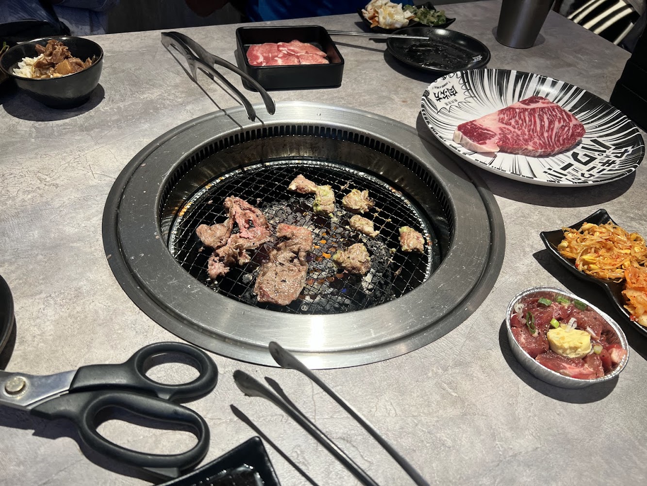 一桌日式燒肉，中央有烤爐正在烤肉，周圍擺放生肉片、沙拉、小菜和剪刀