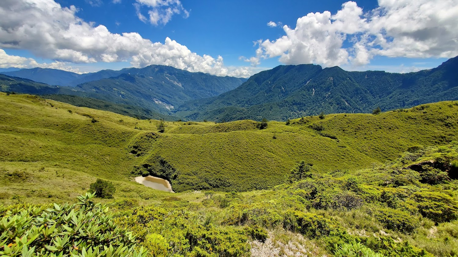這張圖片以壯麗的山脈和翠綠的草原為主,天空藍天白雲,畫面中也有一個小水池,整體景色自然、寧靜且遼闊,沒有明顯浮水印。