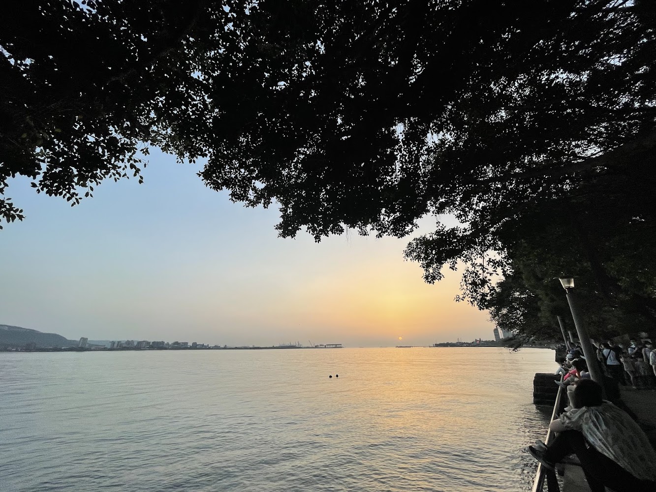 河岸日落景色，民眾在岸邊觀賞夕陽，遠處可見城市天際線倒影