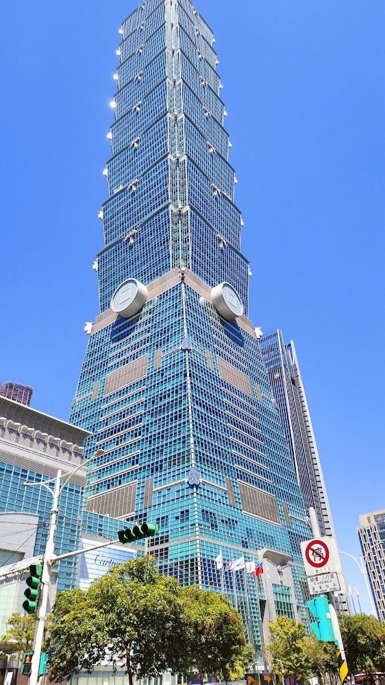 台北101大樓白天外觀，藍綠色玻璃帷幕明顯，前方有樹木與號誌。