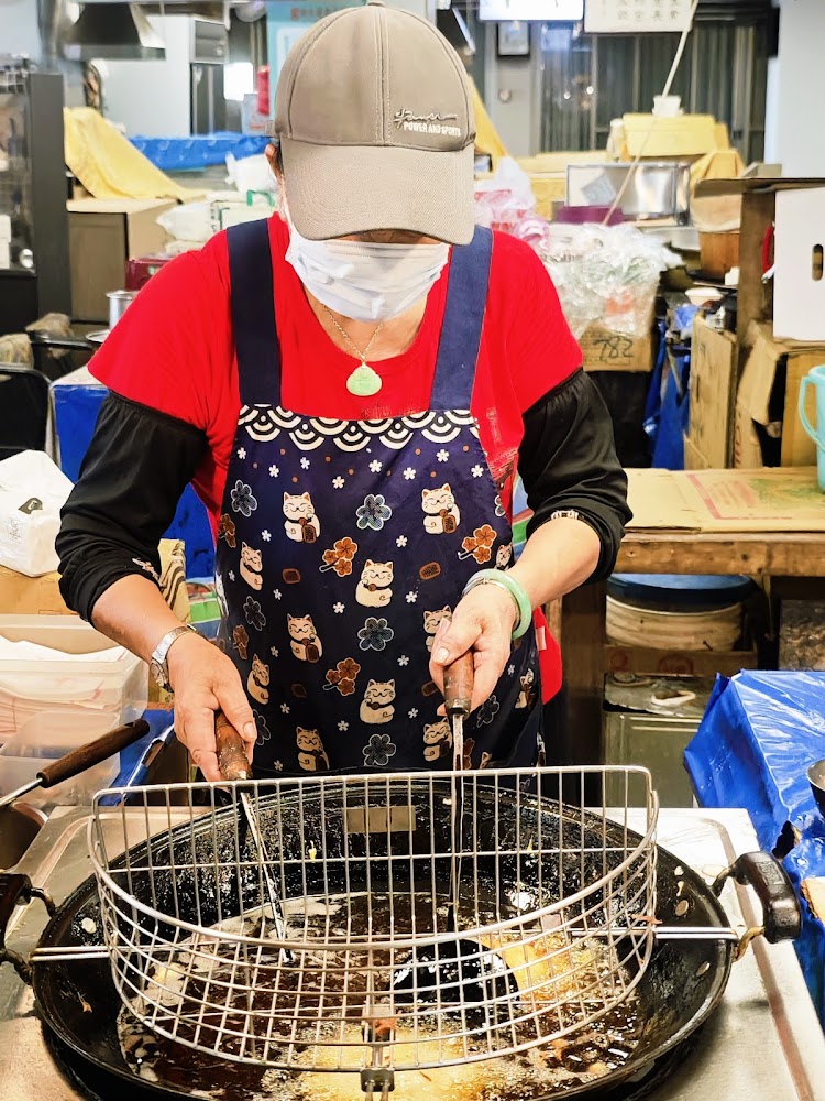 這張圖片主要展示一位穿著貓咪圍裙的攤販阿姨，在市場裡操作油鍋現炸食物，專心炸製美食，現場氣氛溫馨樸實。