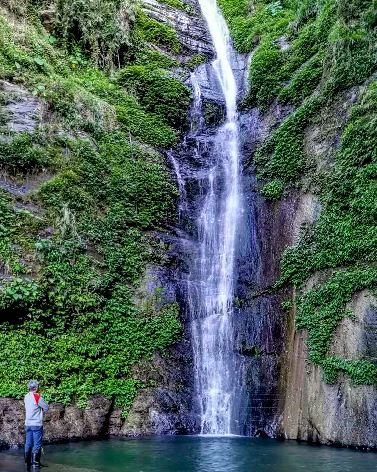 遊客站在烏來瀑布底部仰望，兩側峭壁布滿綠植，水氣瀰漫
