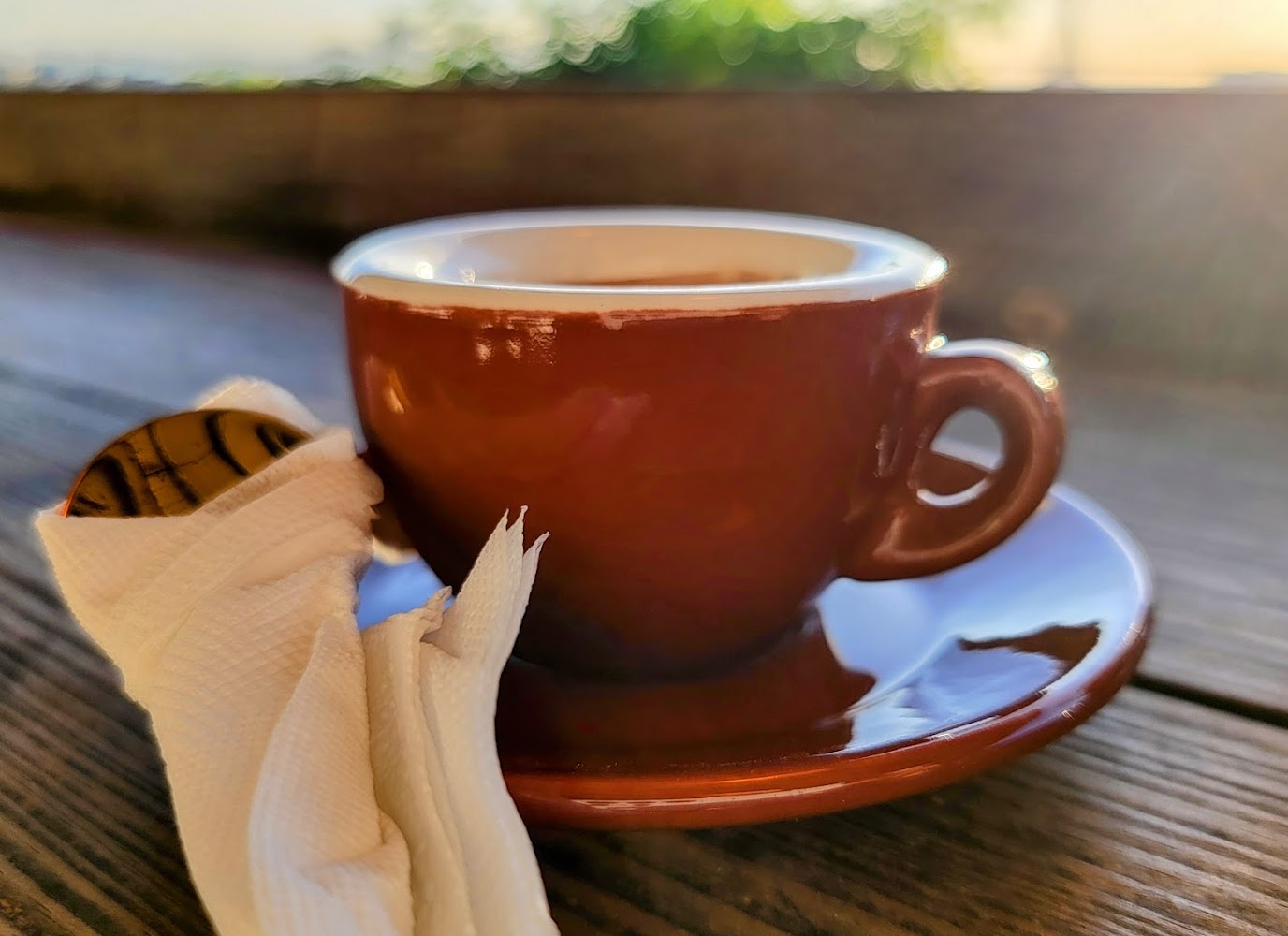 這張圖片的主題是一杯棕色的咖啡杯，咖啡杯放在有木紋的桌面上，旁邊有用紙巾包裹的湯匙，陽光灑落整體溫馨舒適。