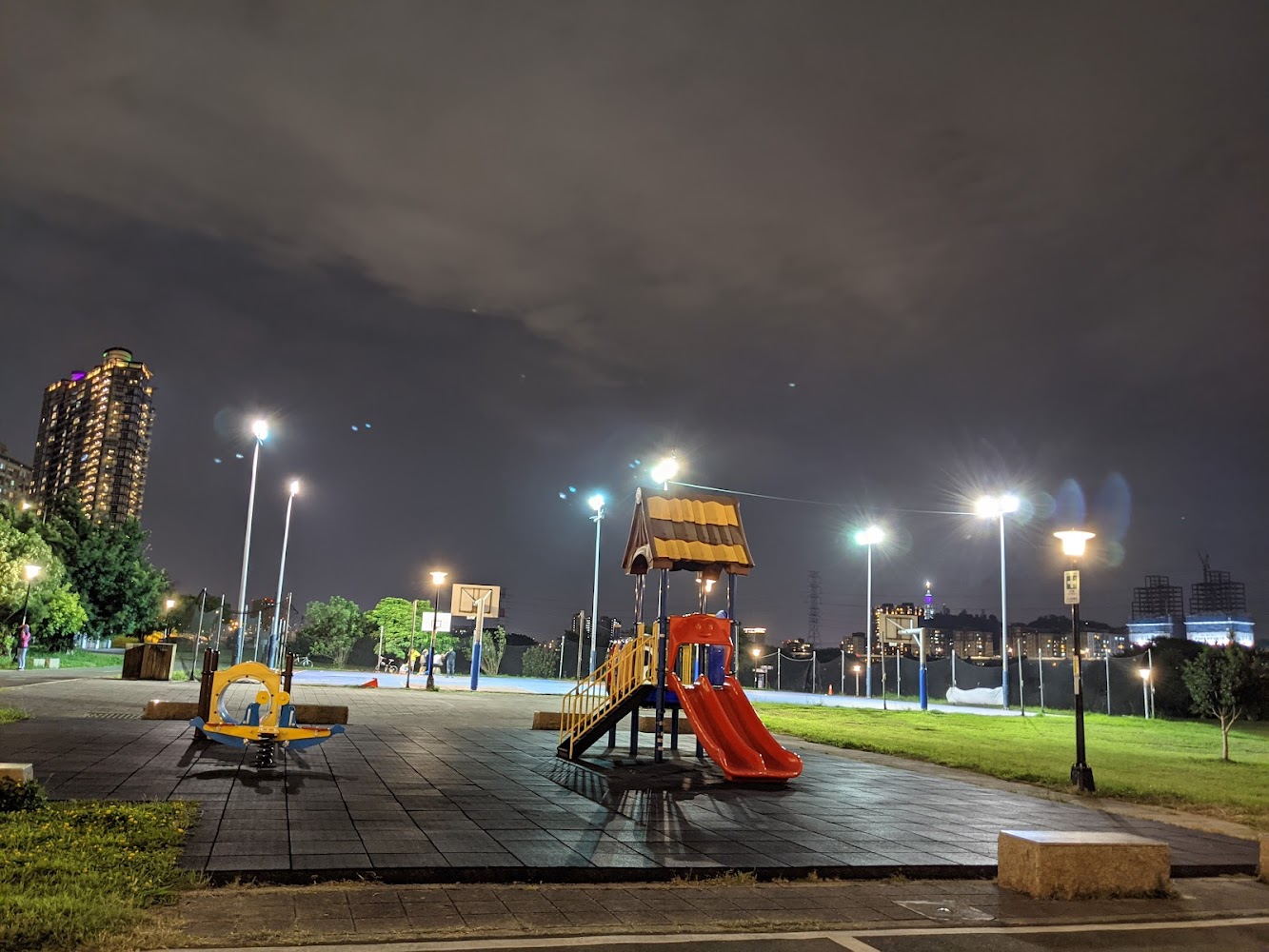 夜晚公園有溜滑梯和搖搖馬，遠處可見籃球場