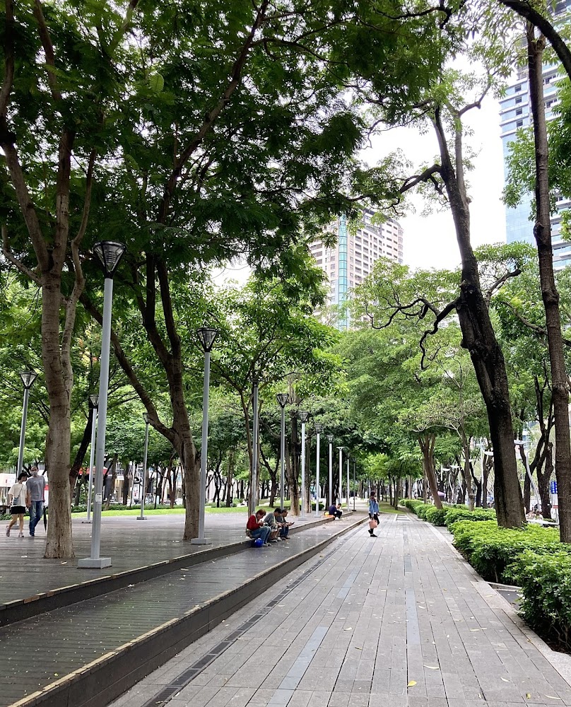 都市中的綠意公園，有整齊樹木、步道與長椅，背景可見高樓
