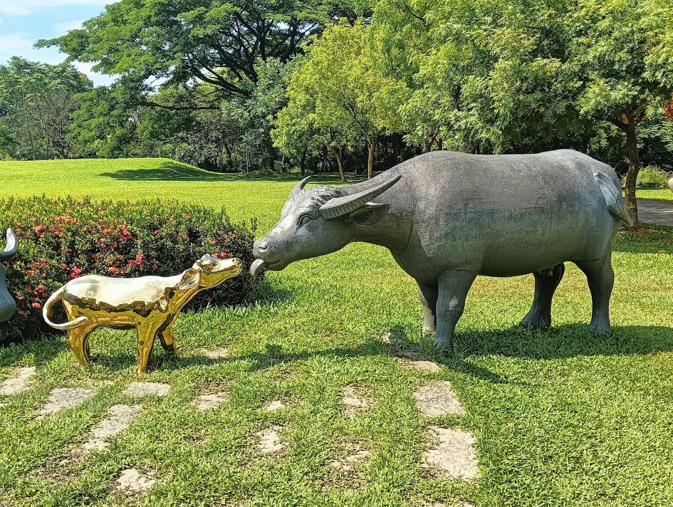 這張圖片展示了一座戶外公園，中央有兩座牛的雕像，其中一隻是金色小牛，另一隻為灰色大牛，它們互相靠近，背景有綠樹與草地。