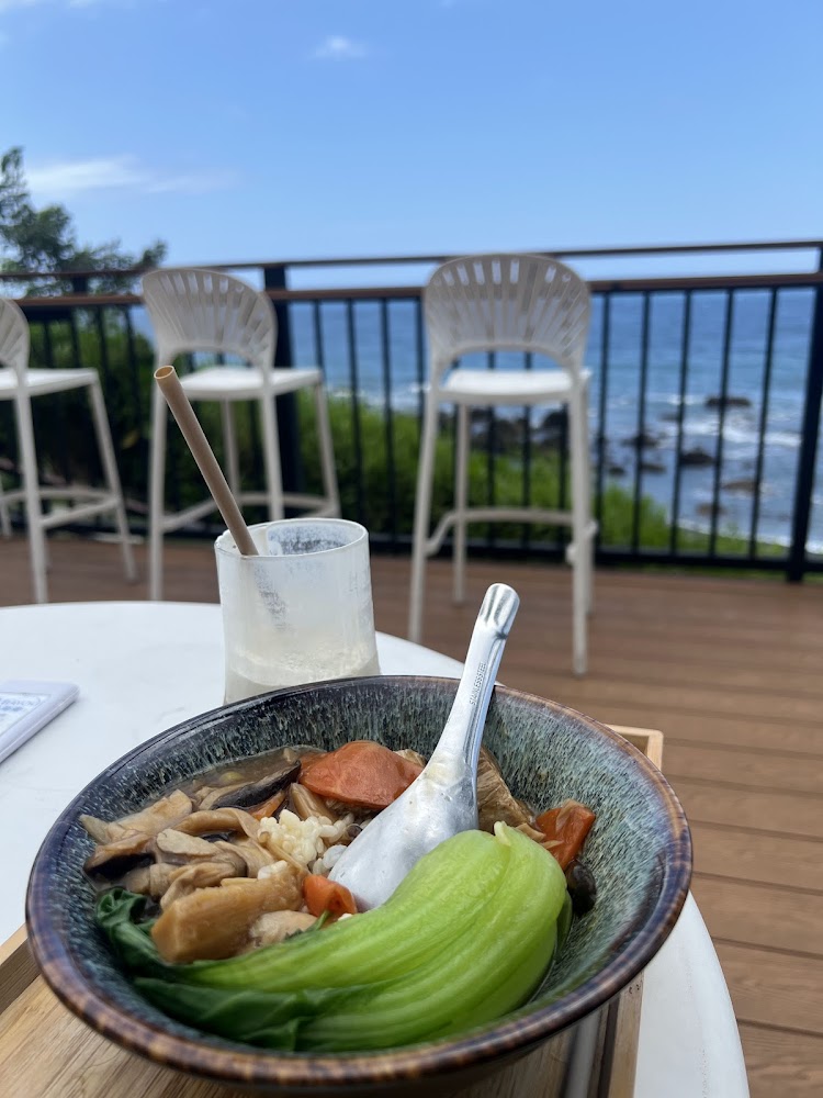 一碗豐富的蔬菜麵，旁邊有一杯飲料，背景是戶外的椅子和大海