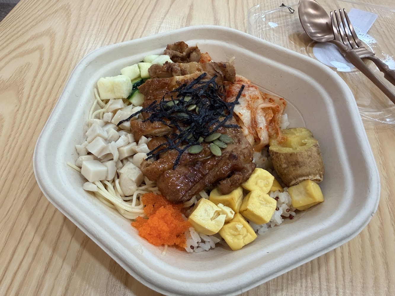 這張圖片展示了一碗豐富的便當，內有烤雞肉、豆腐丁、炒蛋、甜薯、泡菜、小黃瓜、海苔絲及魚卵，底層則舖有白飯或糙米飯，營養均衡且色彩繽紛。