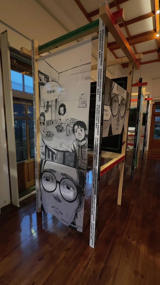 這張圖片展示一個展覽空間,中央懸掛著大型漫畫風格的畫板,上面有家庭聚餐的場景和人物特寫。四周木質結構環繞,空間明亮且現代化。圖片無明顯浮水印。