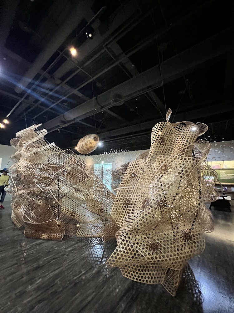 這張圖片展示了一個大型的竹編藝術裝置，懸掛於室內空間中。結構由多片網狀竹編單元組成，造型曲折富有流動感，展現出傳統工藝與現代設計的結合。