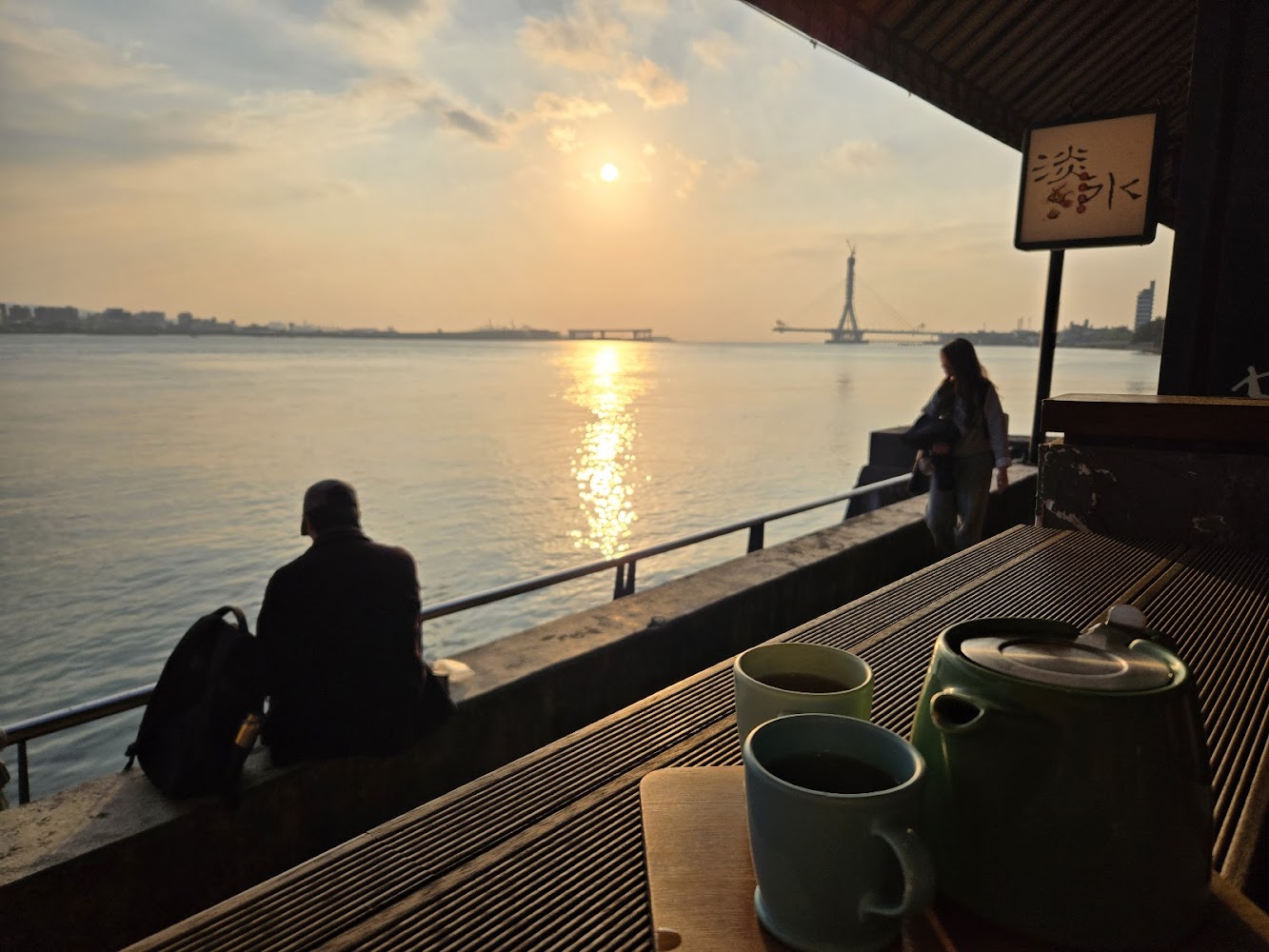 河岸夕陽美景，桌上有茶壺和兩杯茶，遠方有人坐在河邊欣賞風景