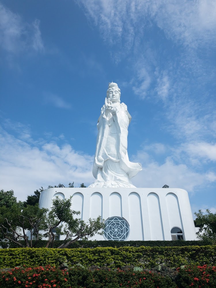 高大的白色觀音佛像矗立在藍天白雲下周圍有綠樹花草