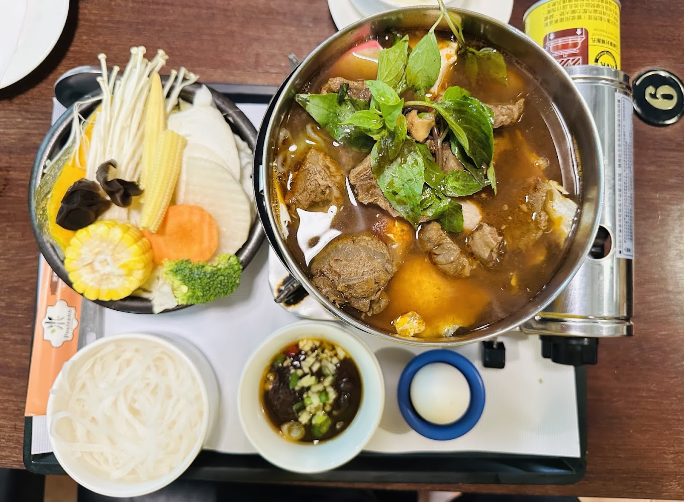 這張圖片主要呈現一份牛肉火鍋套餐，包含豐富的蔬菜拼盤、牛肉湯鍋、白色麵條、沾醬和一顆生雞蛋，配色豐富、擺盤精美。