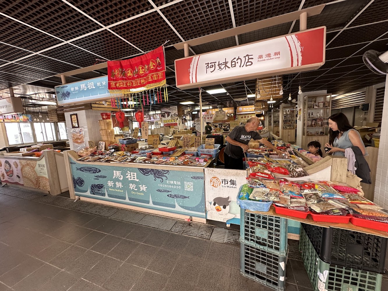 這張圖片是一個傳統市場的攤位，主要販售新鮮和各式各樣的乾貨海產，像是魚乾、蝦米、魷魚等。畫面中有販售者與顧客互動，環境充滿生活氣息。