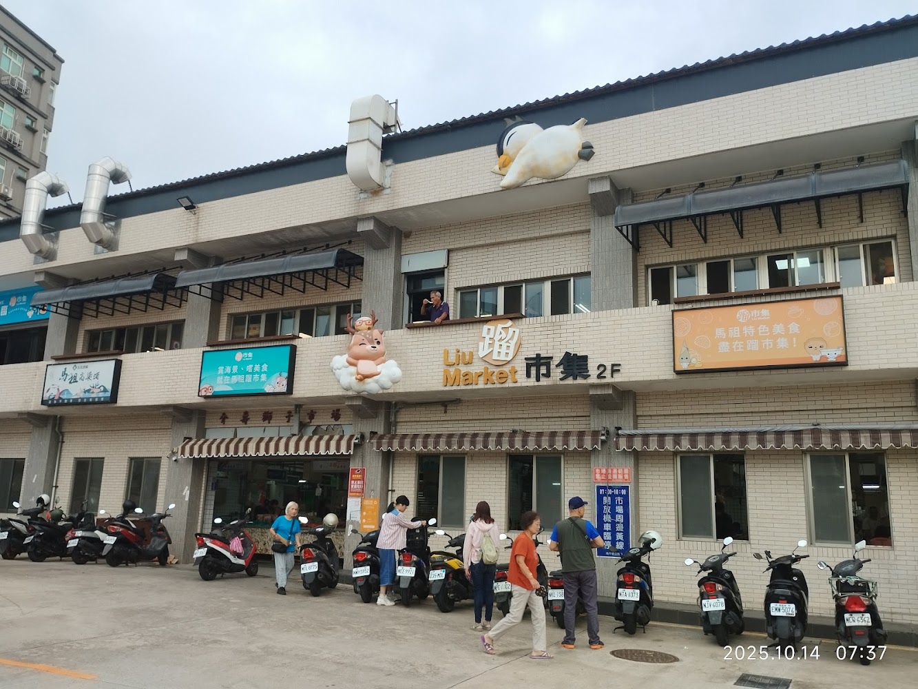 這張圖片主要顯示一棟二層樓市場建築，外牆有可愛動物裝飾，騎樓下停放著多輛機車，有多位民眾在市場前活動，顯示熱鬧的市集氛圍。