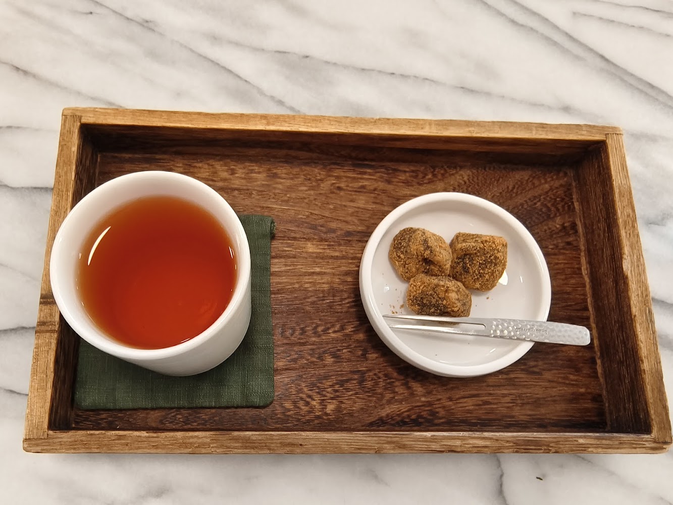 這張圖片展示了一杯熱茶與三塊點心，分別放在木質托盤上，旁邊還有一把小叉子，整體擺設簡潔雅致。