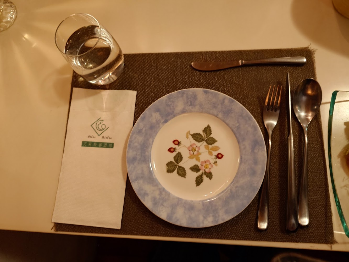 一套餐具擺設，包括花紋瓷盤、刀叉湯匙、水杯以及印有文字的紙巾