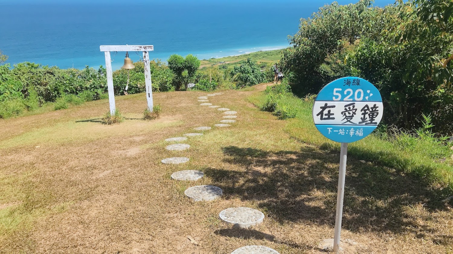 這張圖片展示一片草地，有一條圓形石板步道通往一座白色的愛之鐘，遠方是蔚藍的大海和天空。右側有一個寫著「海線520 在愛鐘 下一站:幸福」的圓形指示牌。