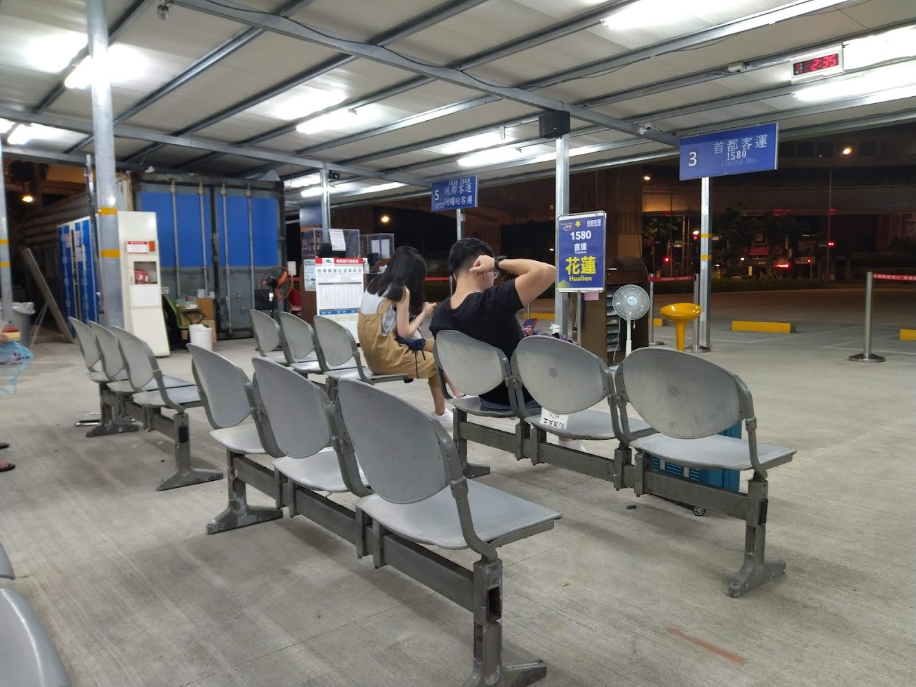 夜晚客運車站，座位區有幾位乘客在等車，旁邊有時刻表和指示牌