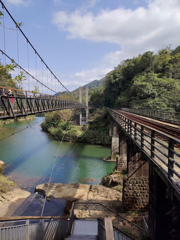 這張圖片主要有一條河流、兩座並行的橋樑（分別為吊橋和鐵道橋），河岸兩側有茂密的綠樹和山丘，天空晴朗，景色優美寧靜。