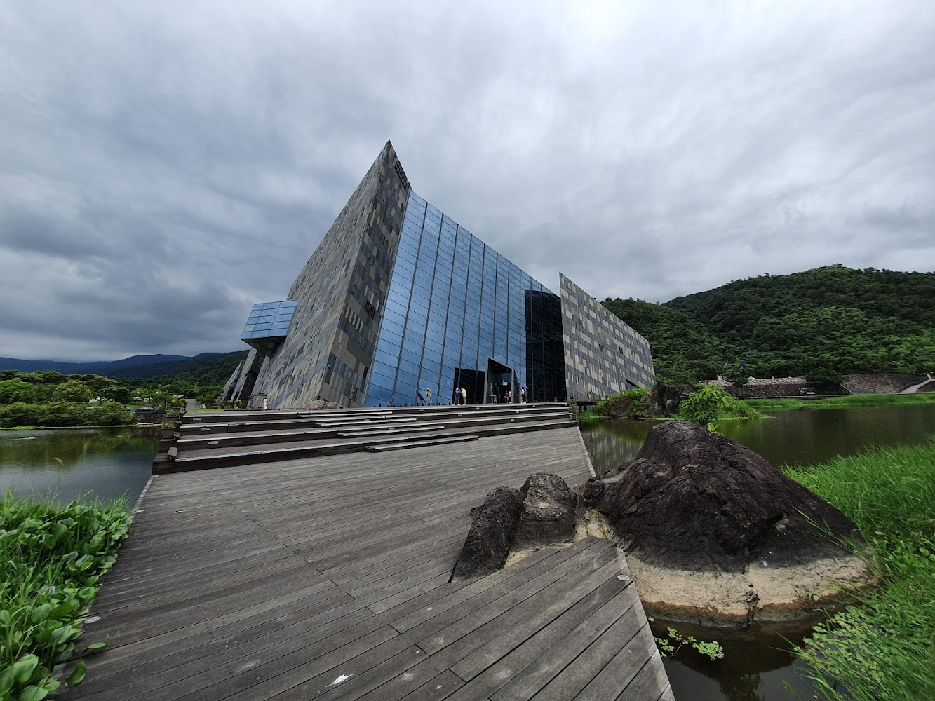造型獨特的現代建築坐落湖畔旁，外觀有大面積玻璃和灰色牆面，四周綠山環繞