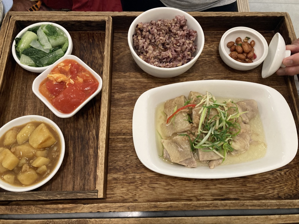 這張圖片是一份中式健康便當，包括白斬雞、紫米飯、燙青菜、番茄炒蛋、滷馬鈴薯以及滷花生，菜色均衡多樣且色彩豐富。