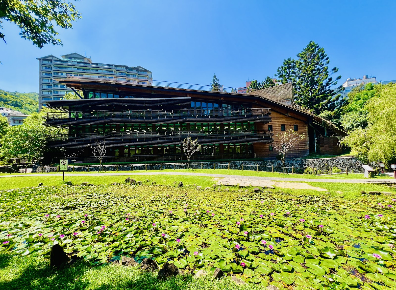 北投圖書館外觀圓弧形木造綠建築，前方荷花池粉紅蓮花盛開，藍天映照