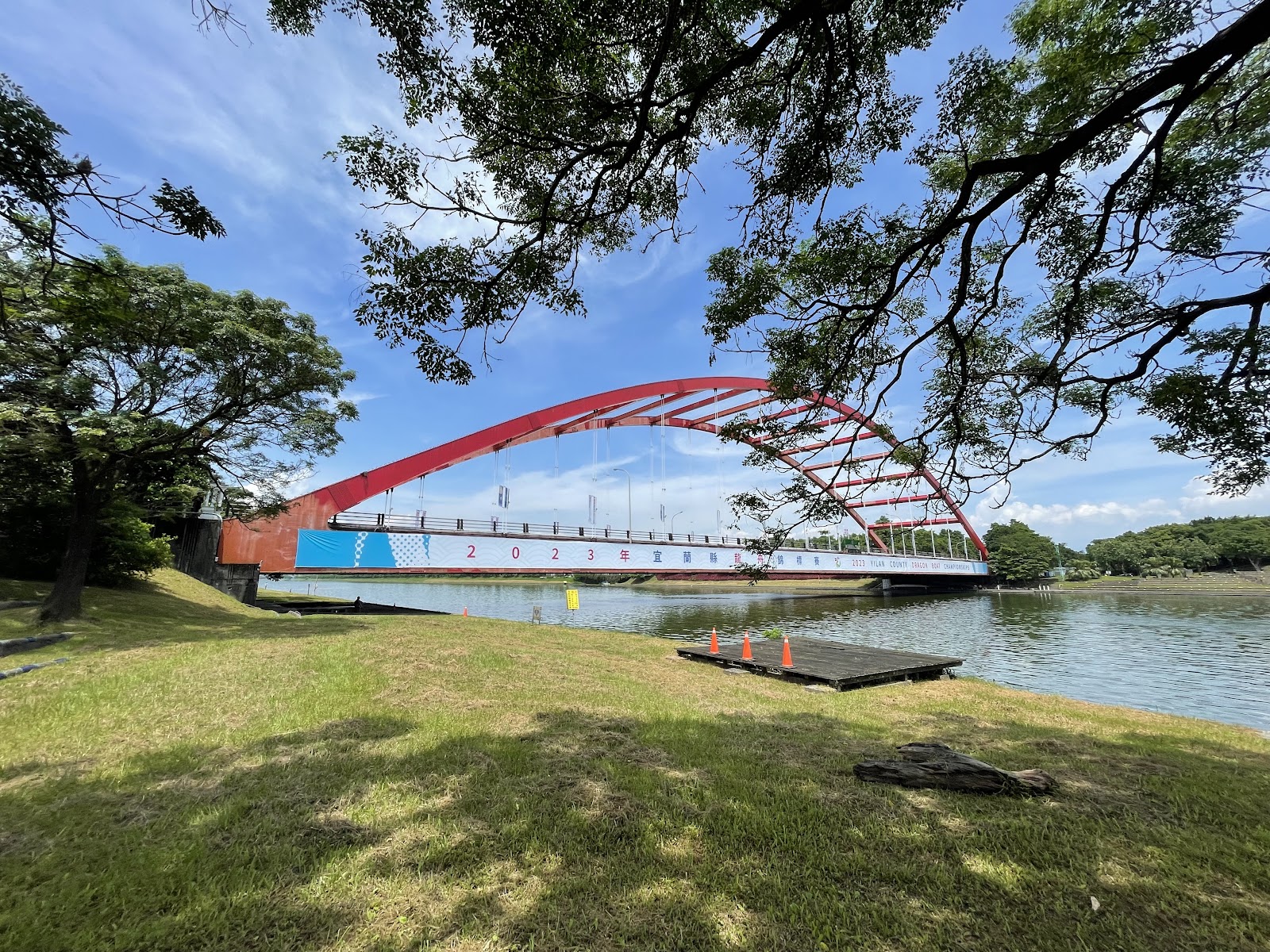 冬山河紅色拱橋從綠蔭樹下框景，2023宜蘭縣國際划水節，河岸草地