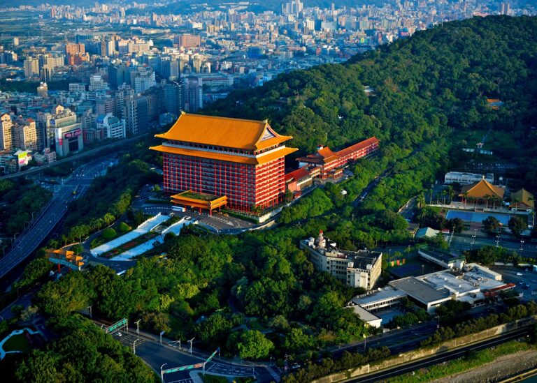 圓山大飯店鳥瞰全景，宮殿式紅色建築配金黃屋頂，雄踞綠色山頭，背景台北市區天際線