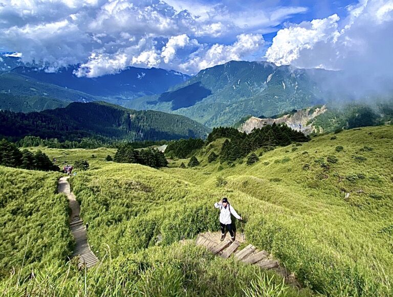 合歡山主峰步道木棧道，登山客攀登翠綠草坡，背景為層疊山脈與藍天白雲