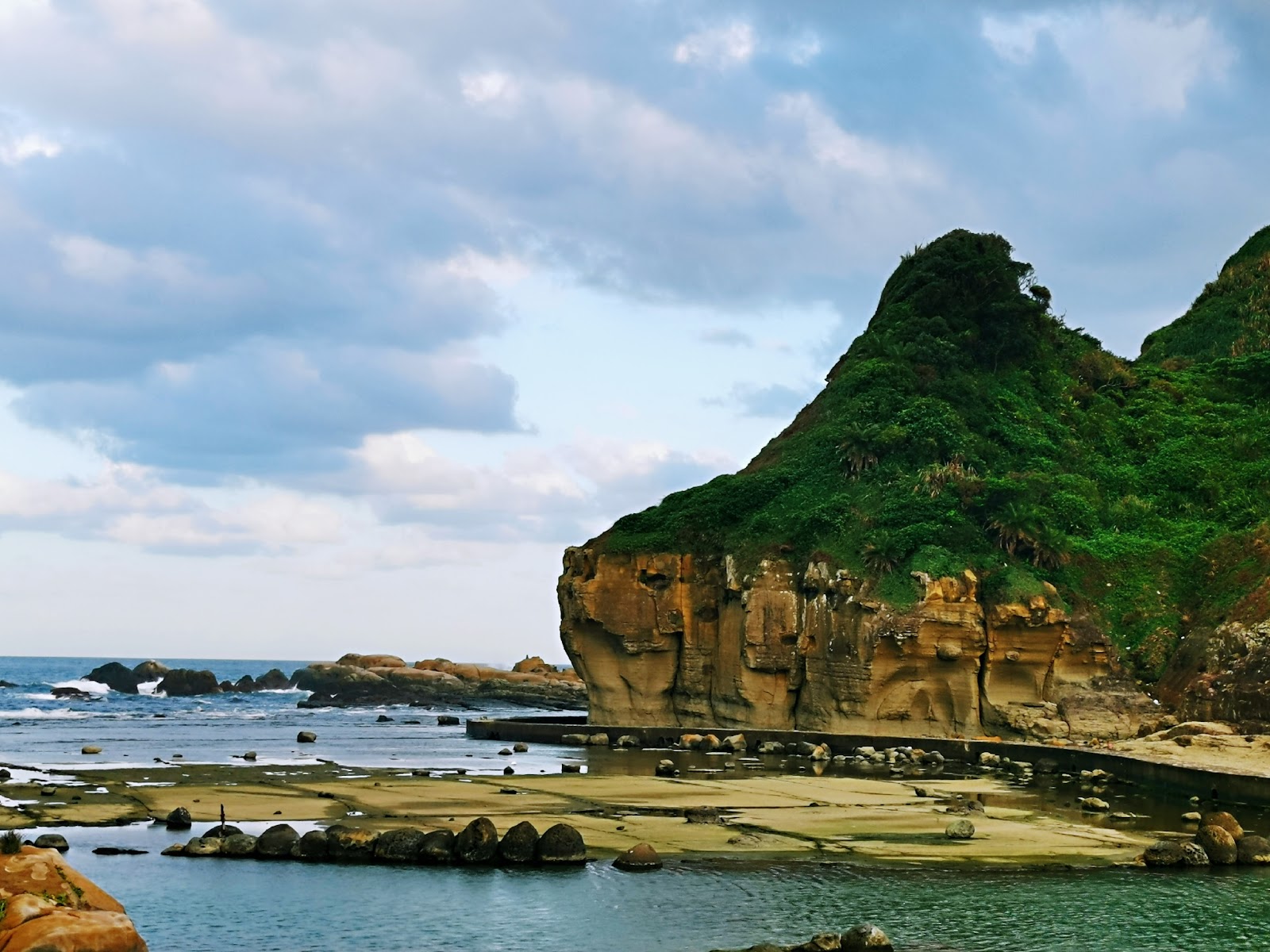 和平島海蝕崖壁近拍,巨大岩石山頭覆蓋翠綠植被,下方黃褐海蝕岩層,前景黃沙小海灘