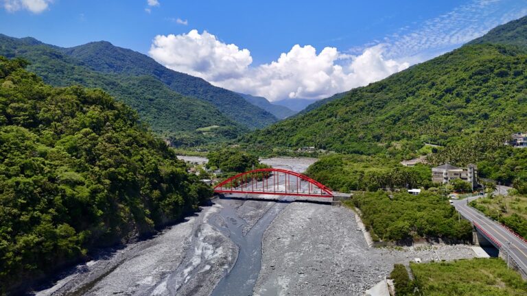 台東金崙溫泉虹橋航拍全景，紅色拱橋橫跨金崙溪礫石河床，兩側青山環繞，藍天白雲