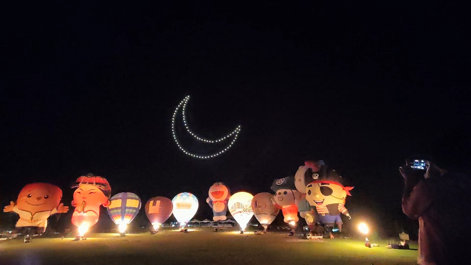 夜空無人機排成月亮圖案，卡通熱氣球在草場發光，觀眾舉手機拍照