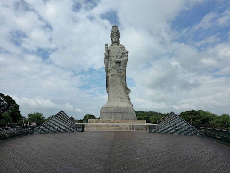 媽祖巨神像正面全景，白色神像高聳入雲，前方木製觀景平台，藍天白雲