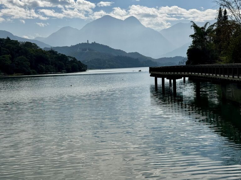 日月潭湖景全景，木棧橋延伸至湖面，遠處慈恩塔矗立山頭，層巒疊翠，藍天白雲