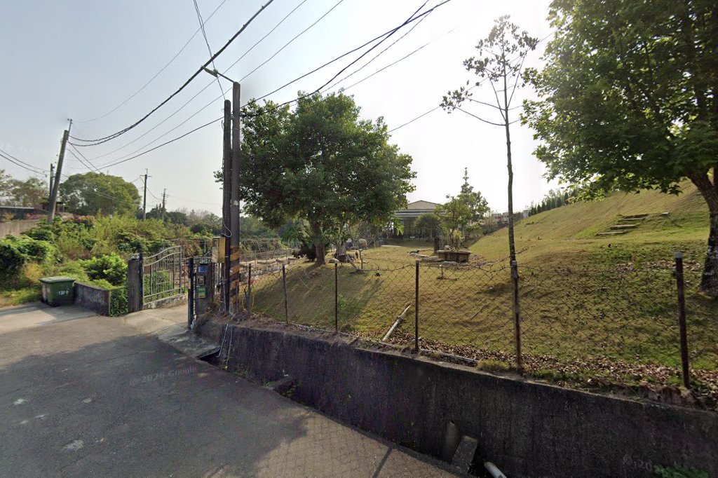 圖片主要顯示一處鄉間道路旁的開放草地，草地上有樹木、圍籬及庭園設施，背景可見住宅建築，天氣晴朗。圖片左側有垃圾桶與鐵門。未見明顯浮水印。